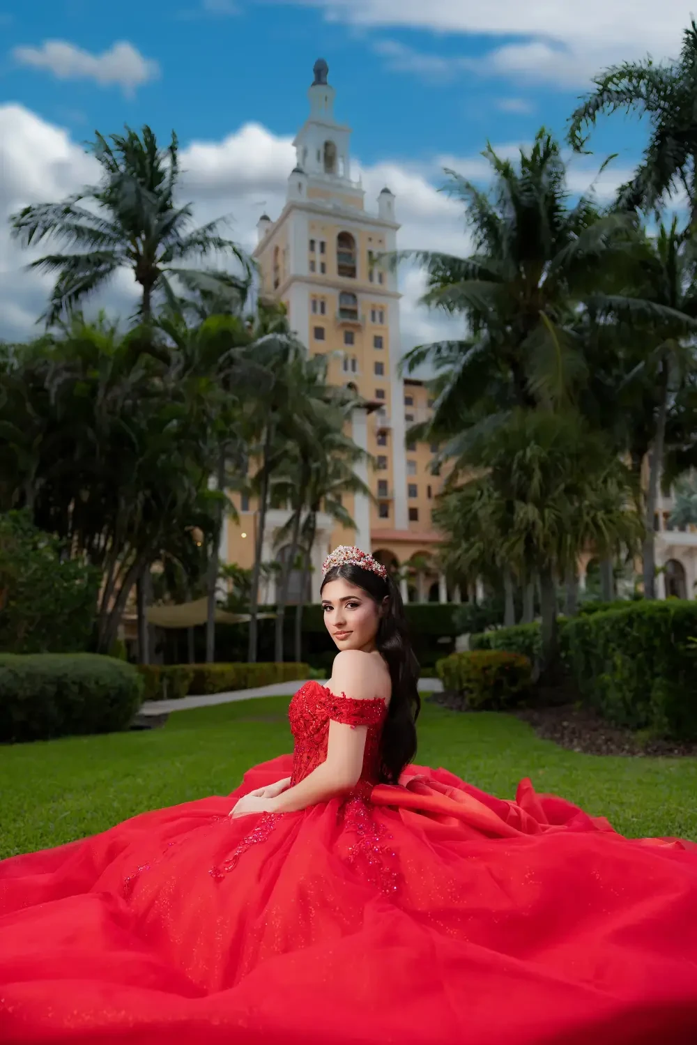 A young woman in a red ball gown sits in a lush garden with palm trees, facing a large building in the background. She wears a tiara and looks serene.