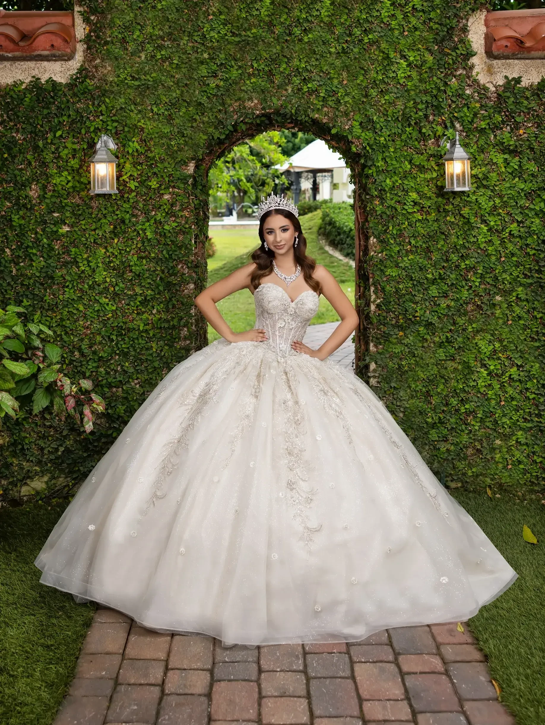 A woman in a luxurious white ball gown stands elegantly in front of a stone archway covered in green foliage, with lanterns hanging on either side.