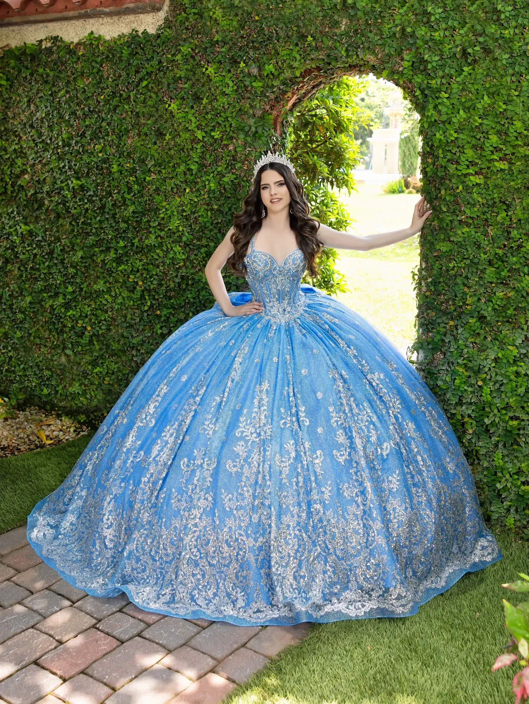 A young woman in a sparkling blue ball gown stands in front of a lush green hedge, framed by an archway. The gown has intricate designs and a full skirt, and she is wearing a tiara. Sunlight filters through the trees in the background.
