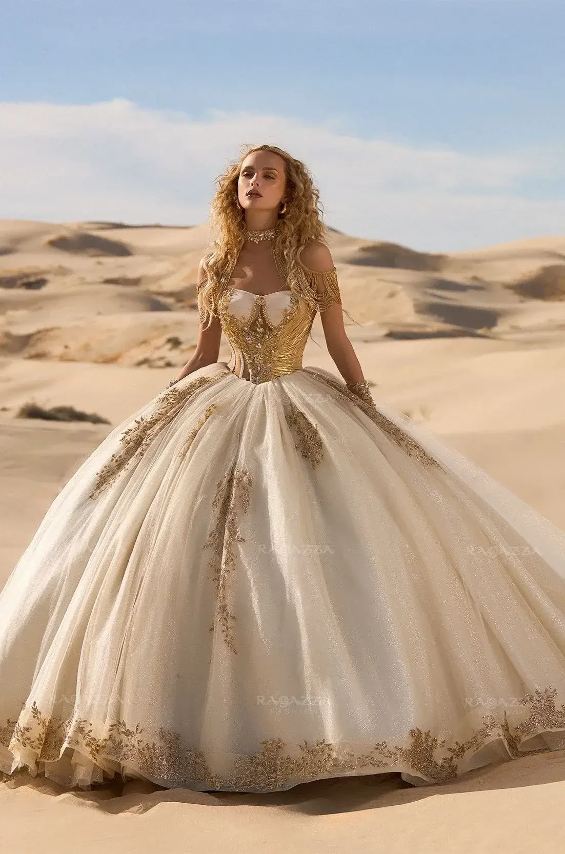 A woman in a flowing, gold-embroidered gown stands in a sunlit desert. Her curly hair and the vast sandy landscape evoke a fairytale elegance.