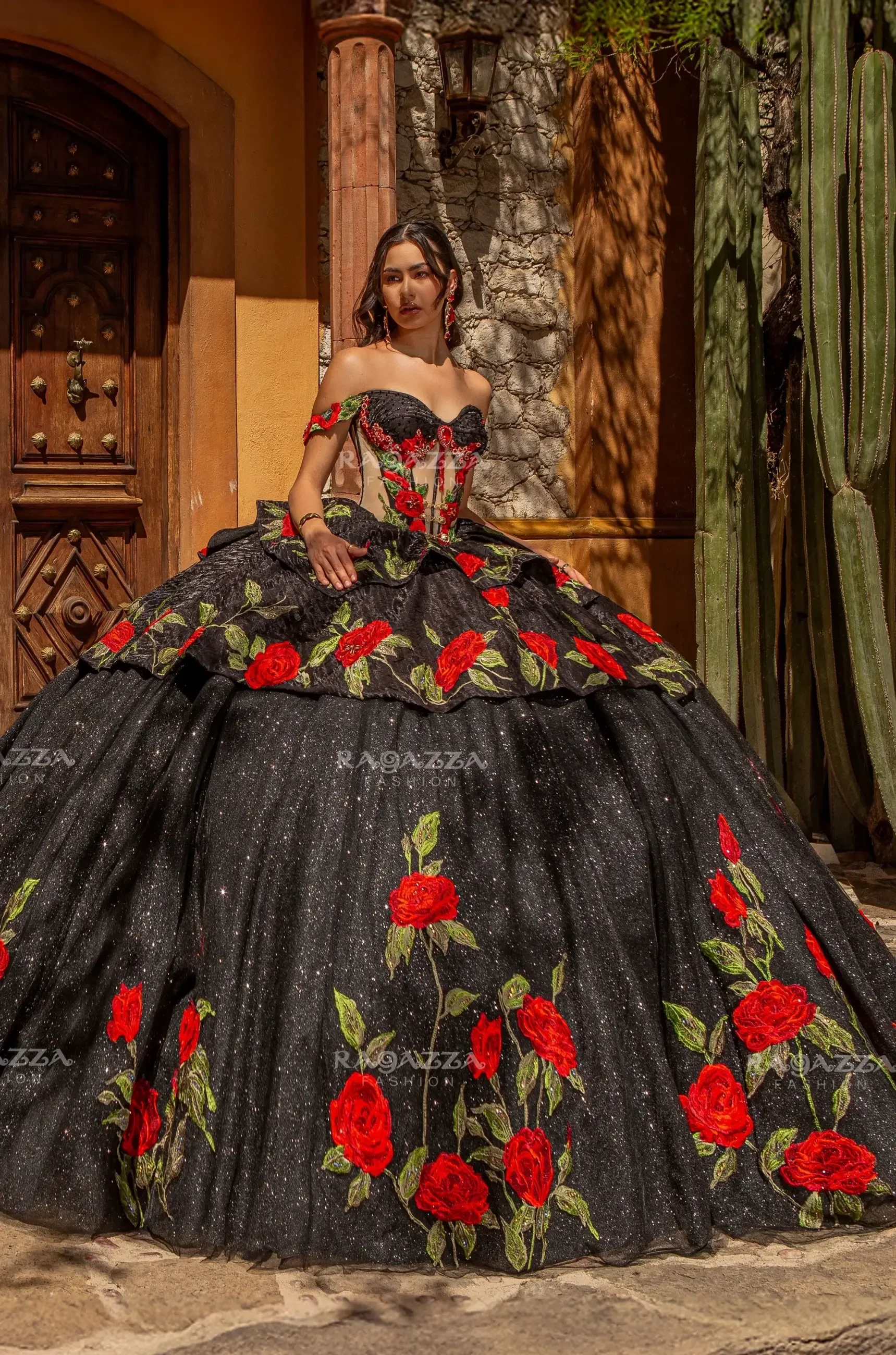 A woman in a strapless black ball gown, adorned with vibrant red roses and green leaves, stands elegantly outside a rustic building with wooden doors and cactus plants.