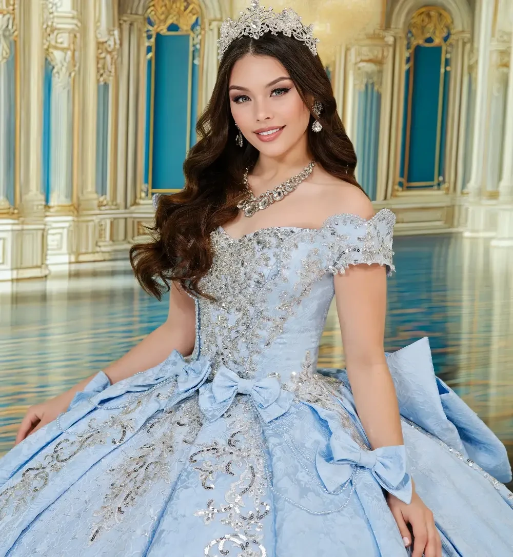 A young woman in a blue ball gown adorned with sequins and bows, wearing a tiara and earrings, smiles elegantly in an ornate palace setting.